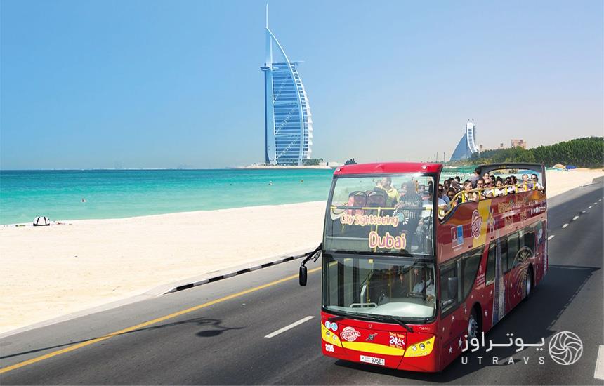 اتوبوس دوطبقه گردشگری قرمزرنگ که با جمعیت مسافران پر شده‌است، درحال عبور از ساحل و تصویر هتل برج‌العرب در پس‌زمینه